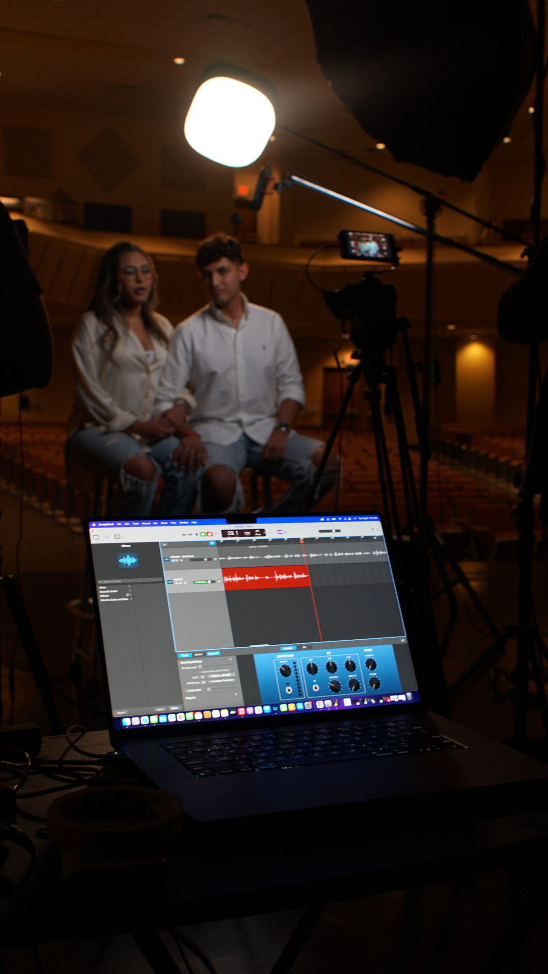 Audio editing laptop in foreground with a two-person interview setup and camera lighting in the background